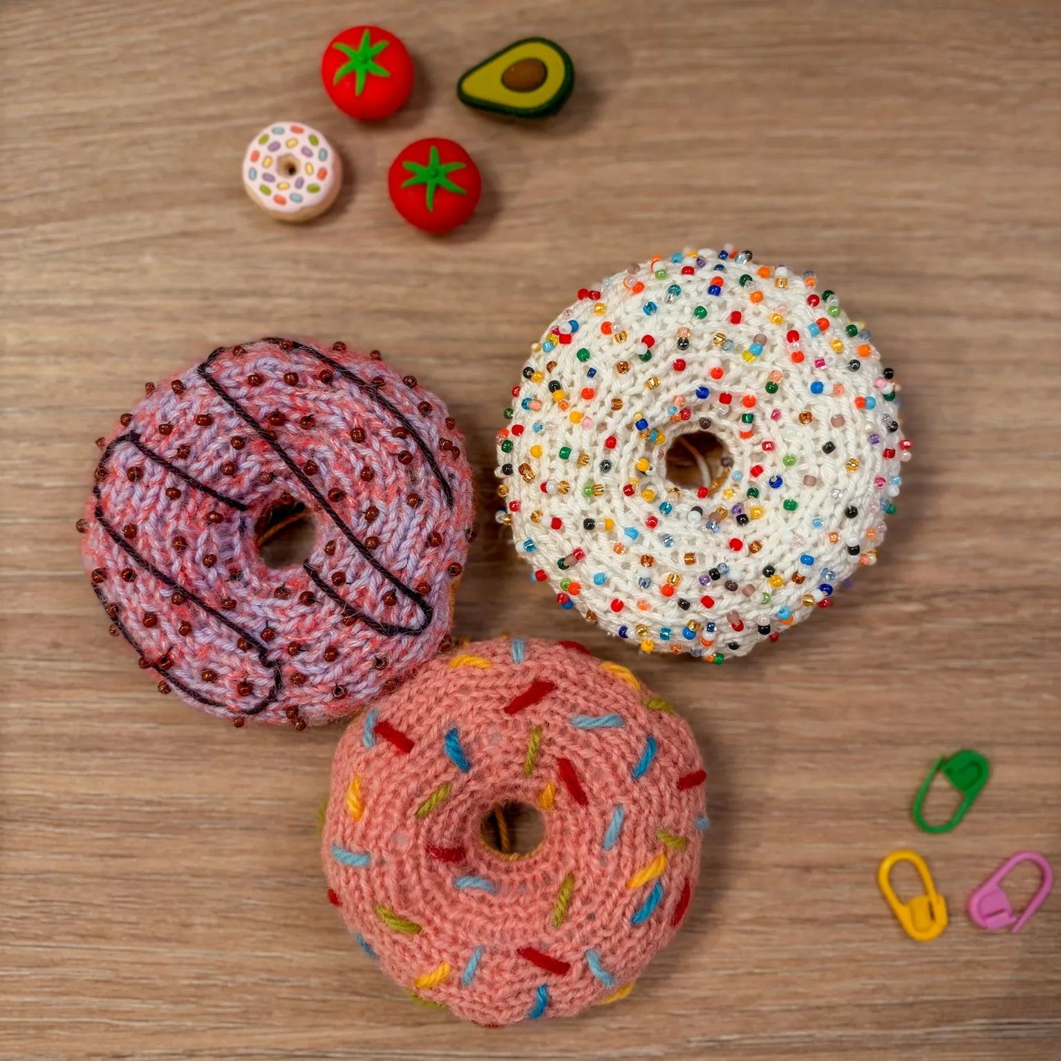 Three knitted donuts on a wooden table: white with bead sprinkles, pink with yarn sprinkles and lilac with a chocolate drizzle